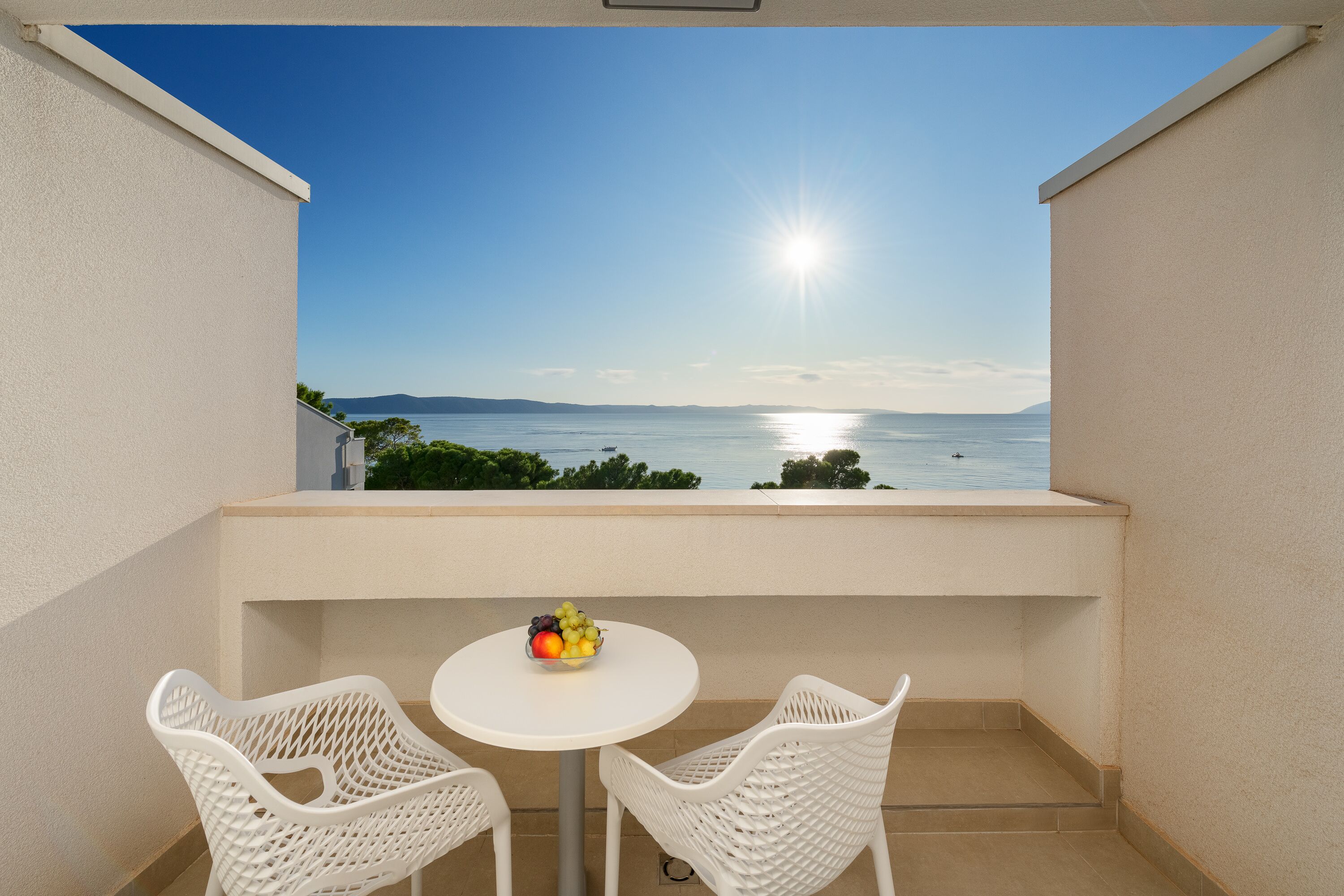 A balcony with two white wicker chairs and a small round table, a bowl of fruit, and a view of the sea and distant mountains under a clear sky.