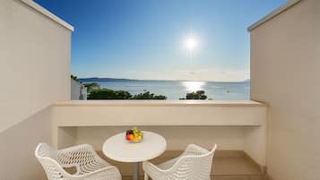 A balcony with two white wicker chairs and a small round table, a bowl of fruit, and a view of the sea and distant mountains under a clear sky.