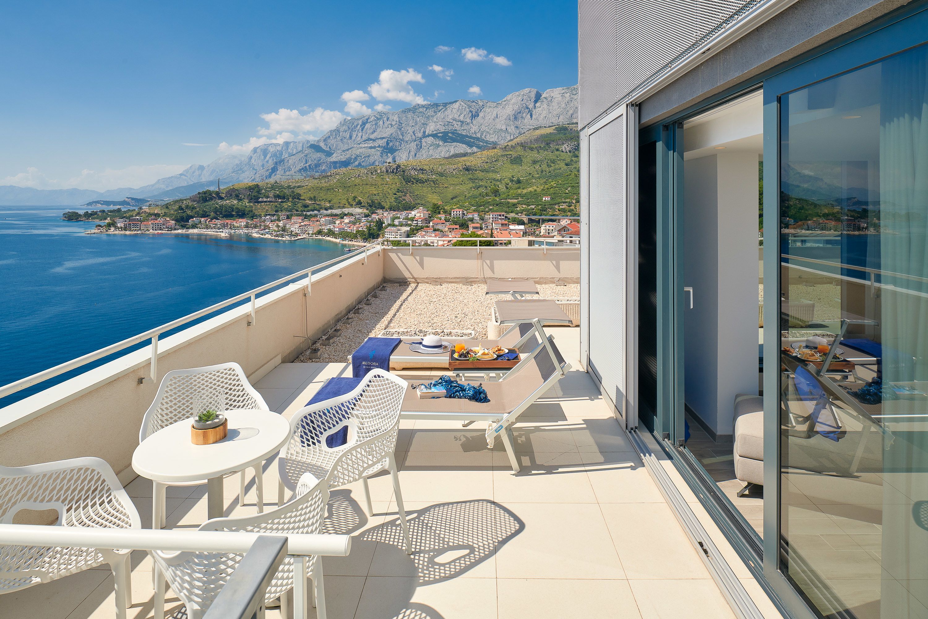 A balcony with white wicker furniture, a small table with a plant, and a view of the sea and mountains.