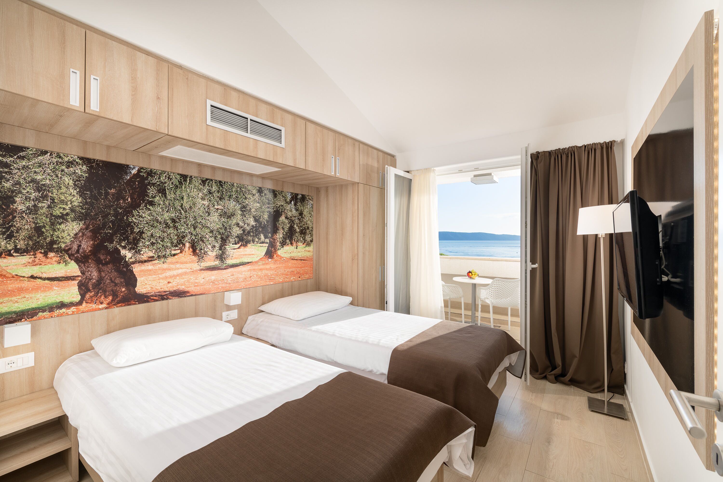 Habitació d'hotel amb dos llits, un quadre gran d'un arbre, televisió i vista al mar.