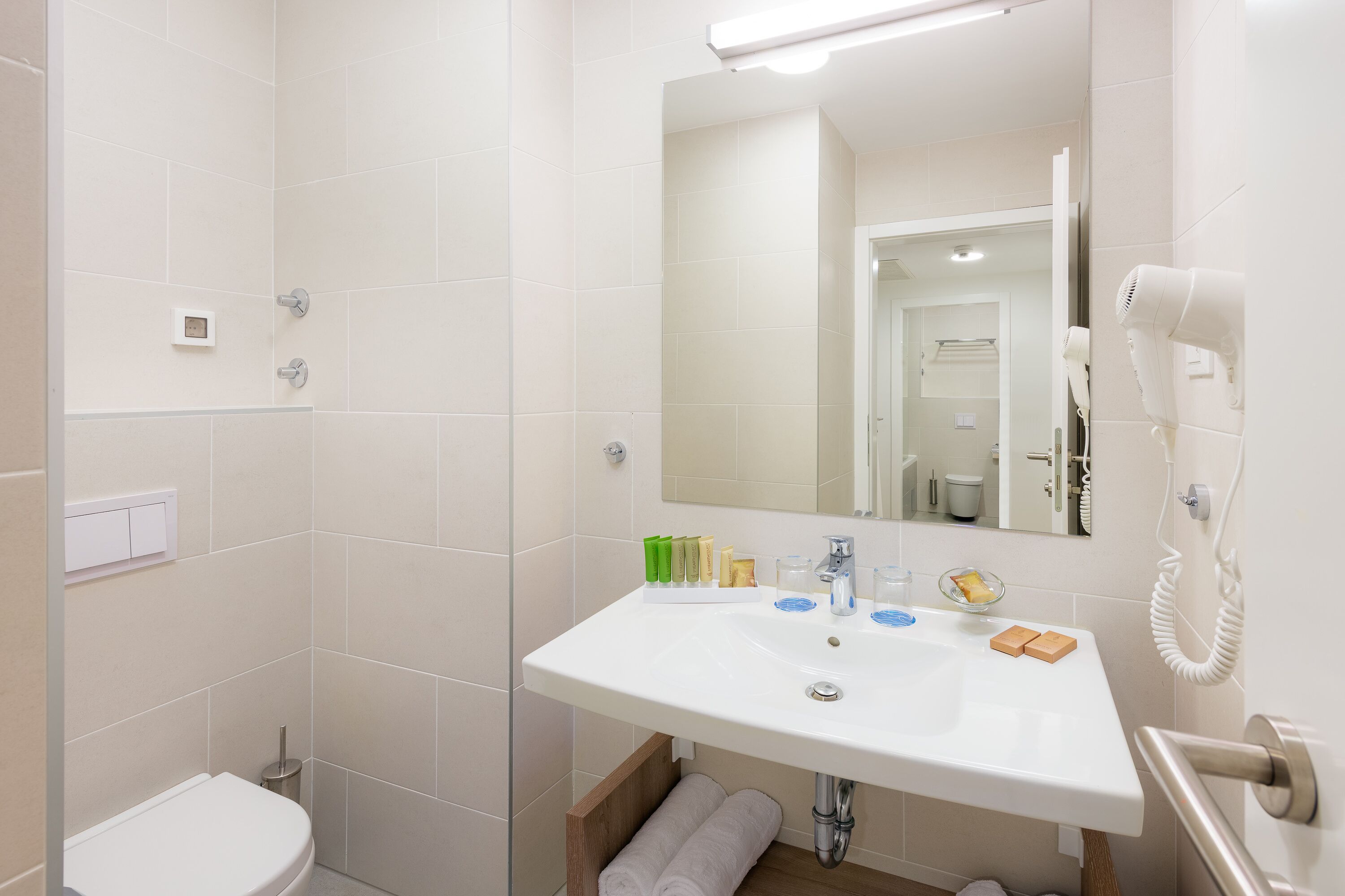 Un baño con lavamanos blanco, espejo y inodoro. Hay artículos de tocador en el lavamanos y un secador de pelo en la pared.