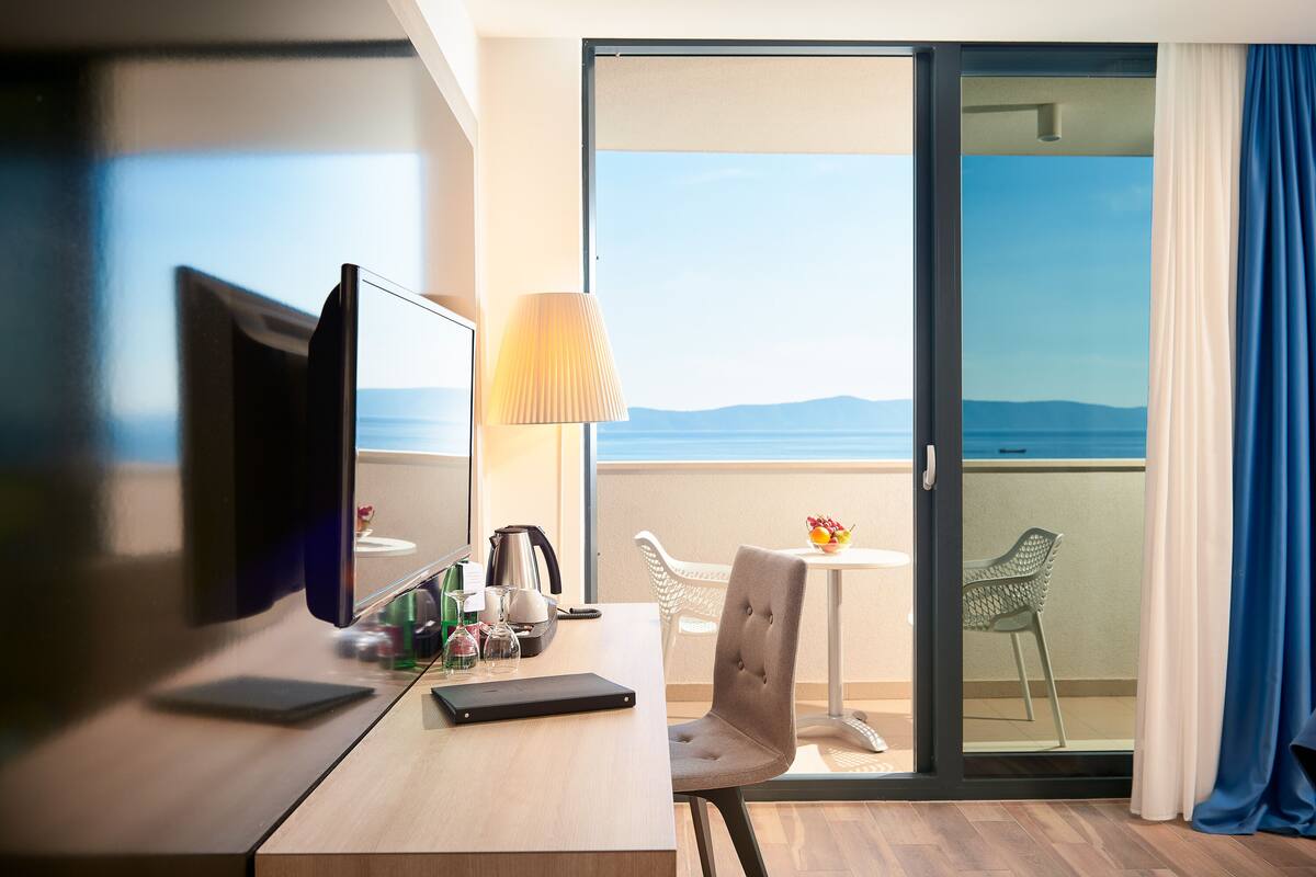 Hotelkamer met een bureau, een laptop, een lamp, een stoel en uitzicht op zee door een schuifdeur.