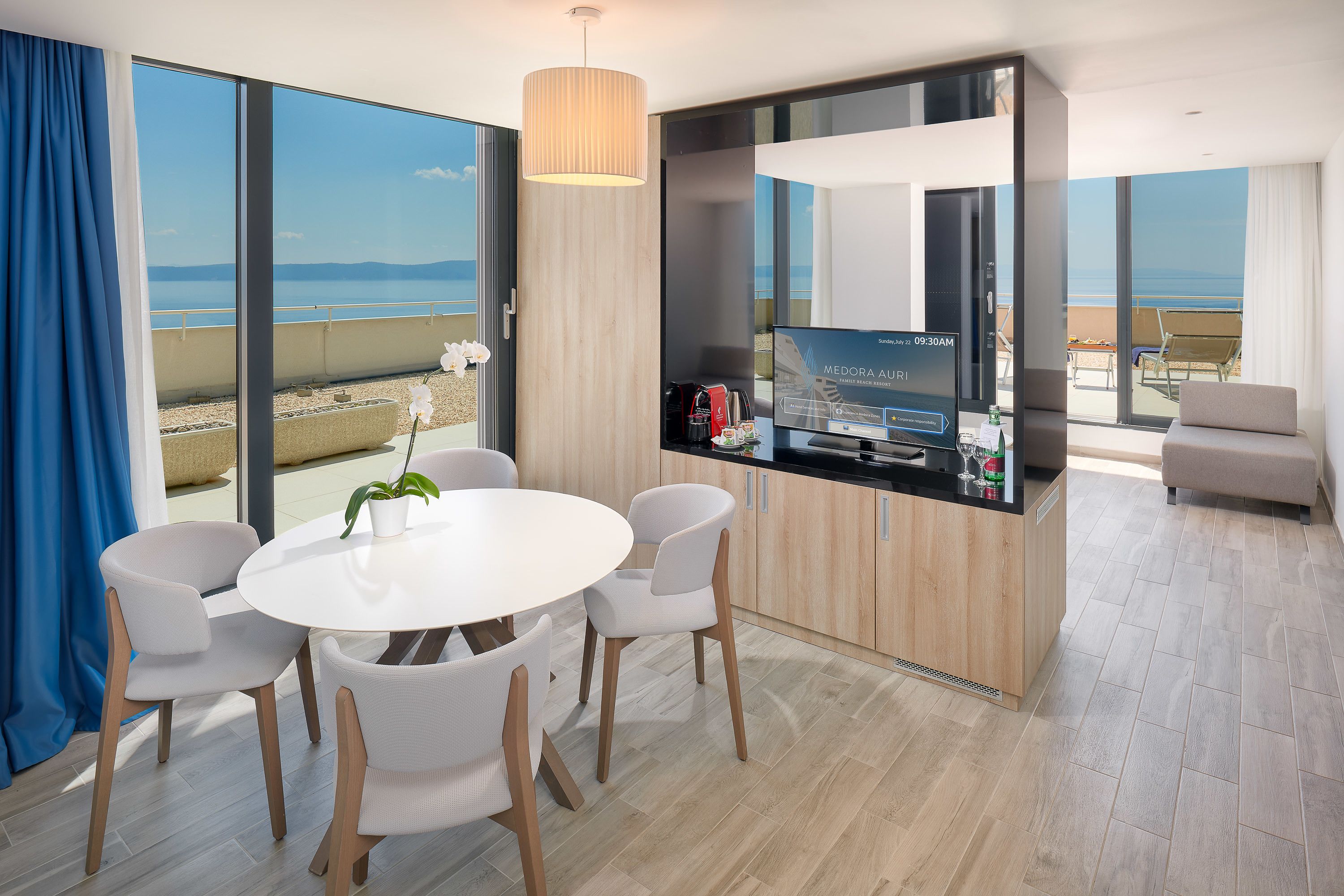A modern hotel room with a dining area, a kitchen counter, and a large window with a view of the beach.