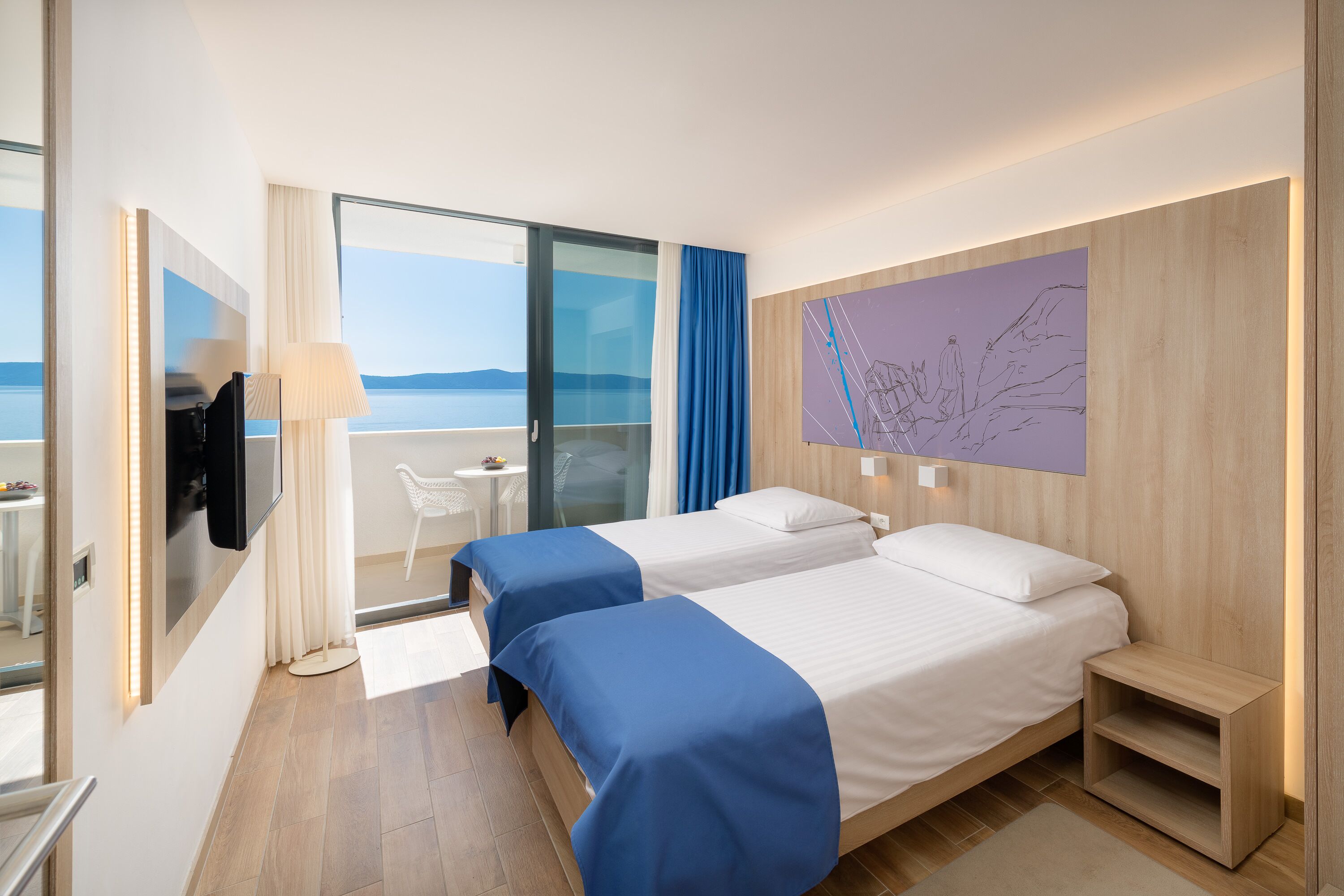 A hotel room with two beds, a TV, a desk, and a view of the sea.