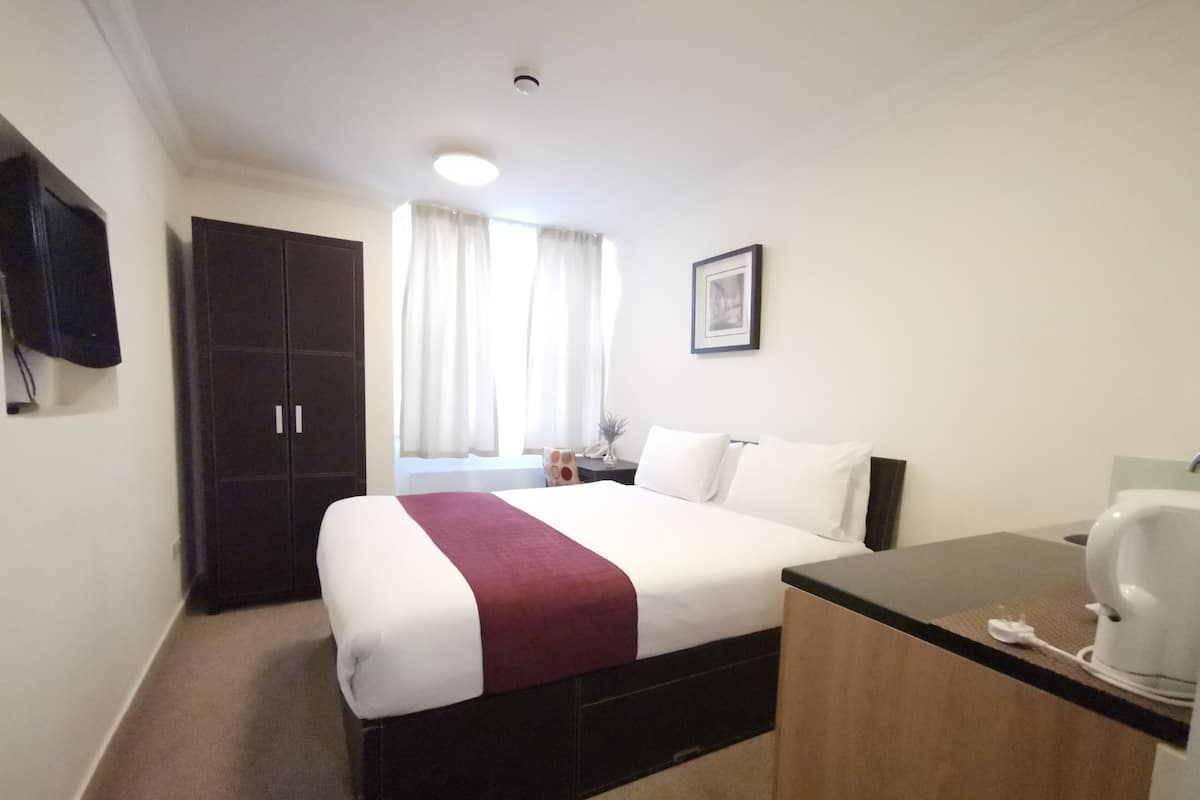 Ein Hotelzimmer mit einem Bett, einem Schreibtisch, einem Stuhl, einem Fenster mit Vorhängen, einem Bild an der Wand und einem Fernseher.