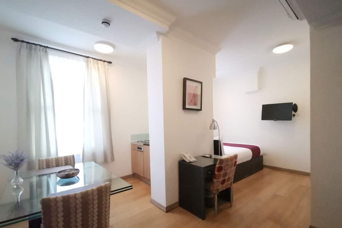 Ein kompaktes Hotelzimmer mit Glastisch, einer kleinen Küchenzeile, einem Bett mit roter Decke, einem Schreibtisch mit Stuhl und einem Wand-Fernseher.