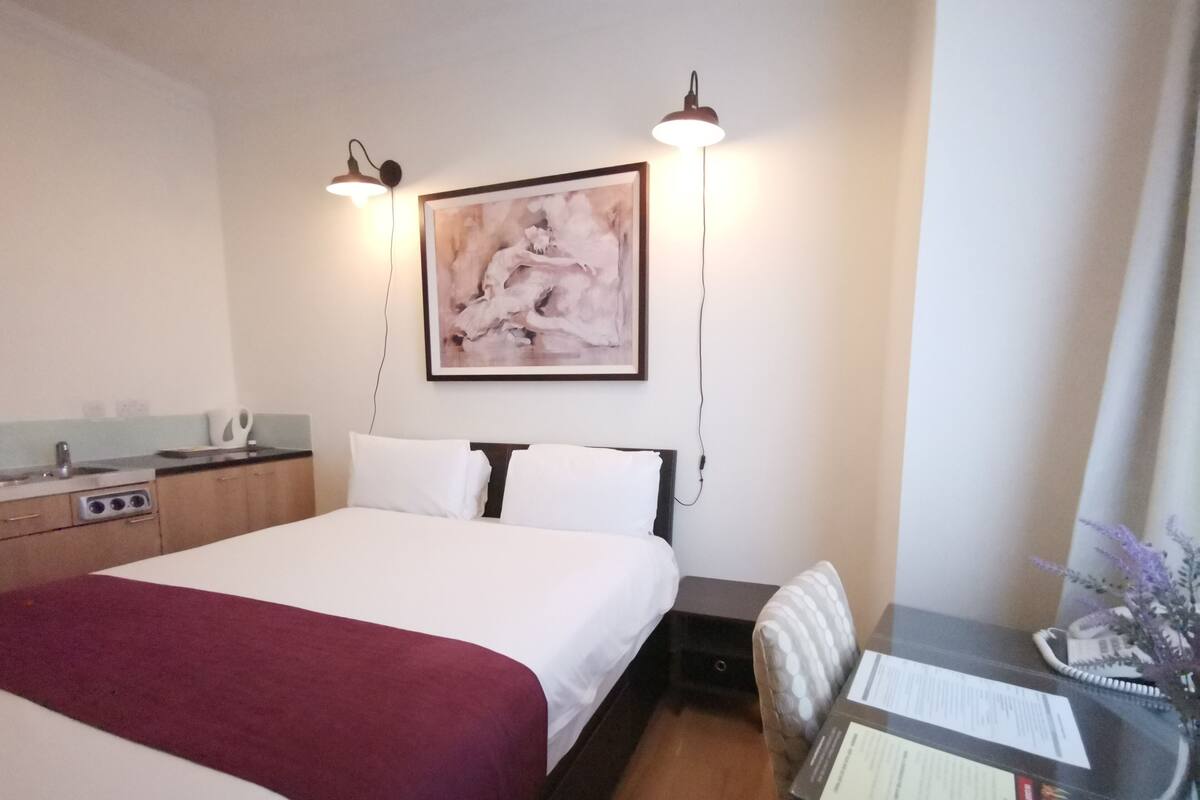 Ein Hotelzimmer mit einem großen Bett, einem Schreibtisch mit Stuhl, einer kleinen Küchenzeile und einem Bild an der Wand.