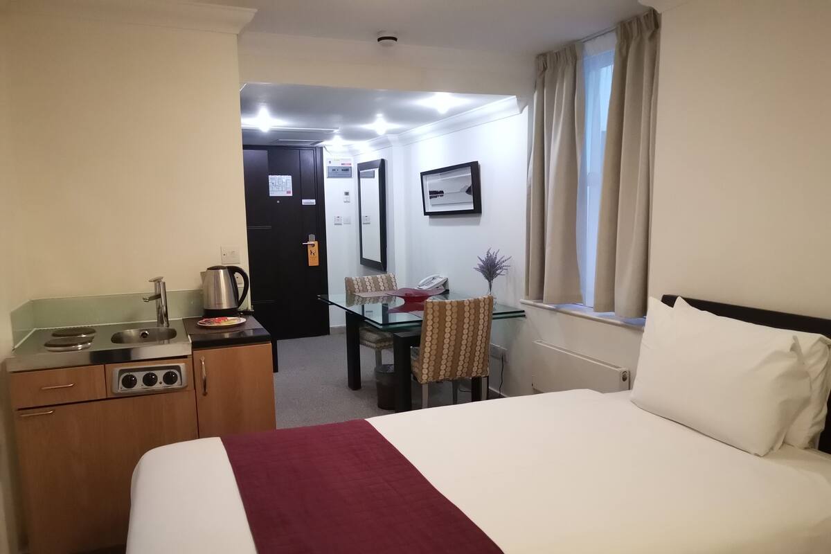 Ein Hotelzimmer mit Bett, Schreibtisch mit Stuhl, einer kleinen Küchenzeile und einem Fenster mit Vorhängen.