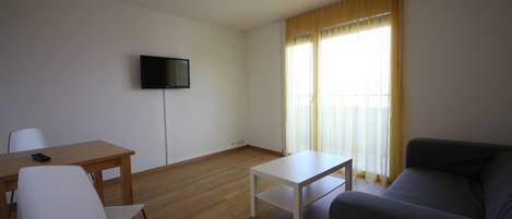 Una sala de estar con piso de madera, una mesa blanca, un sofá gris, una silla blanca y un televisor de pantalla plana montado en la pared.
