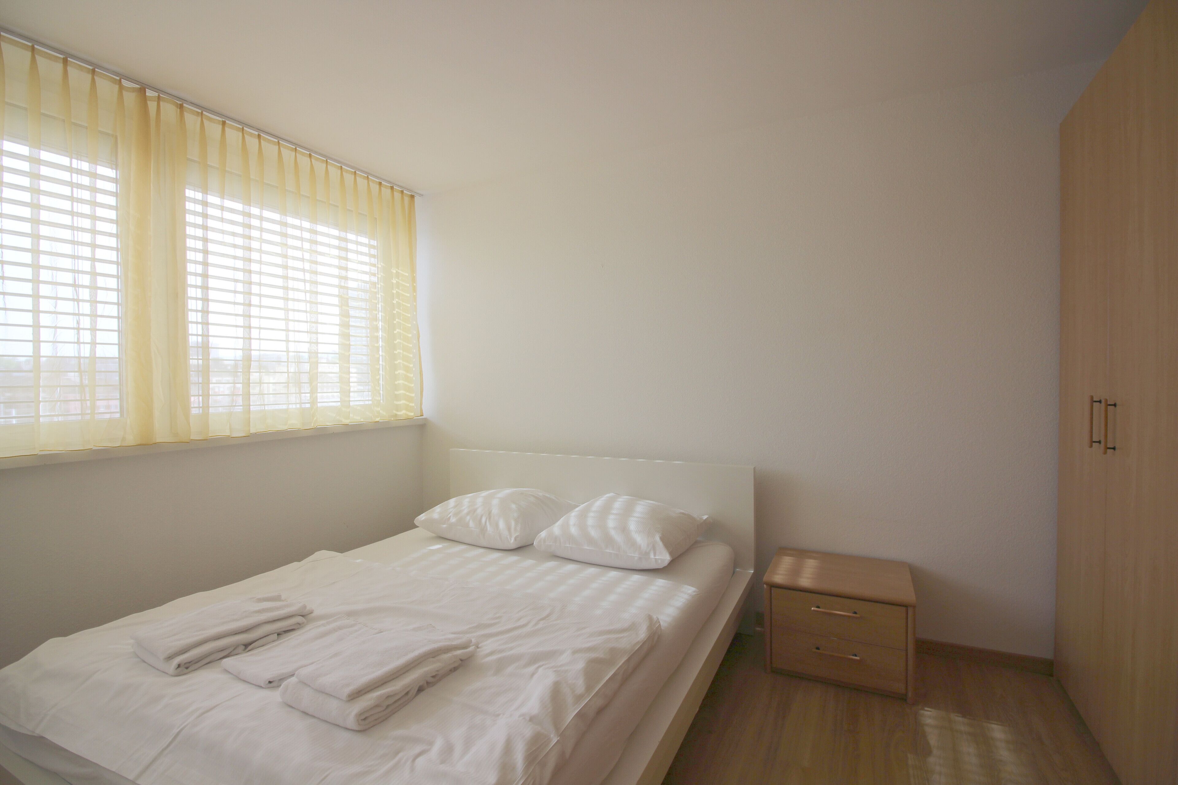 A neatly made bed with pillows and folded towels, a bedside table, and a wardrobe in a simple, well-lit room.