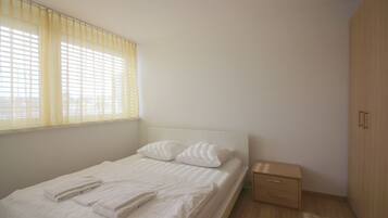 A neatly made bed with pillows and folded towels, a bedside table, and a wardrobe in a simple, well-lit room.