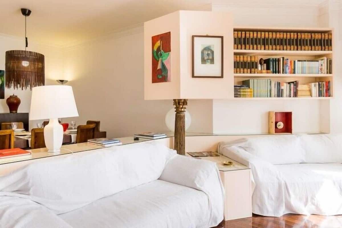 A modern hotel room with a white sofa, a dining table with chairs, a bookshelf, and a hanging lamp.