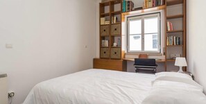 Une chambre avec un lit simple, un bureau en bois avec une chaise, une bibliothèque et une fenêtre.