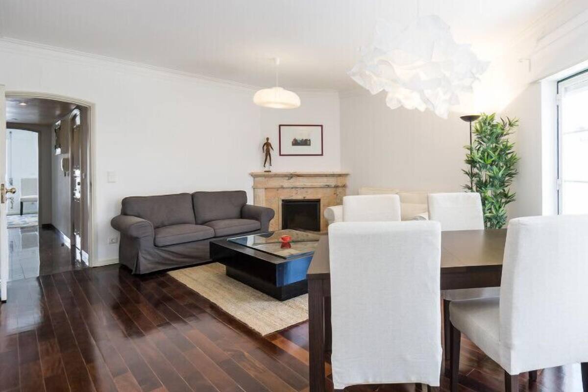 A living room with a sofa, coffee table, dining table with chairs, a fireplace, and a large window with curtains.