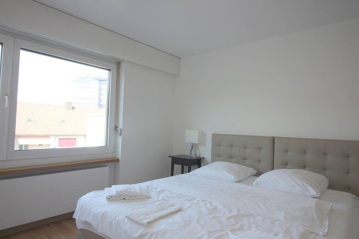 Een hotelkamer met een groot raam, een bed met witte lakens, een nachtkastje met een lamp en een hoofdbord.