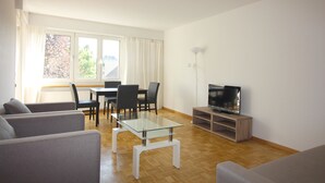 Een moderne woonkamer met een glazen salontafel, grijze banken, een eethoek met stoelen, een televisie op een houten dressoir en een groot raam met gordijnen.