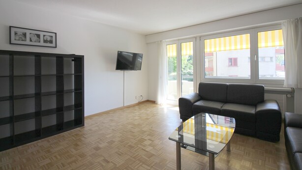 Ein Wohnzimmer mit einem schwarzen Sofa, einem Glastisch, einem an der Wand montierten Fernseher und großen Fenstern mit gelben Vorhängen.