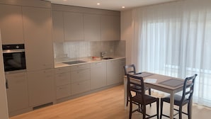 Een moderne keuken met ingebouwde apparatuur, een eethoek met een tafel en stoelen, en grote ramen met dunne gordijnen.