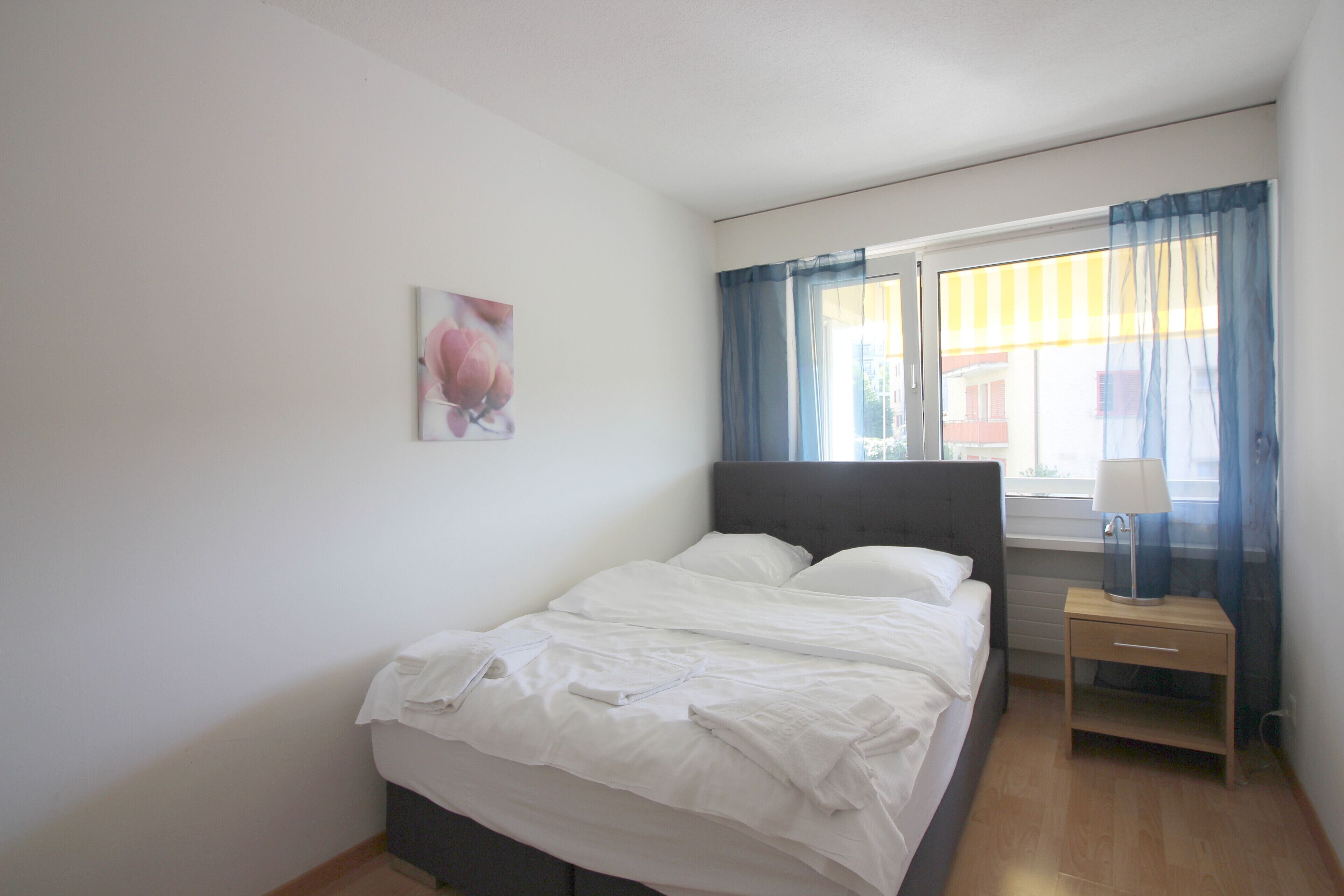 Un dormitori amb llit, una nitzeta, una làmpada, una finestra amb cortines i un quadre penjat a la paret.