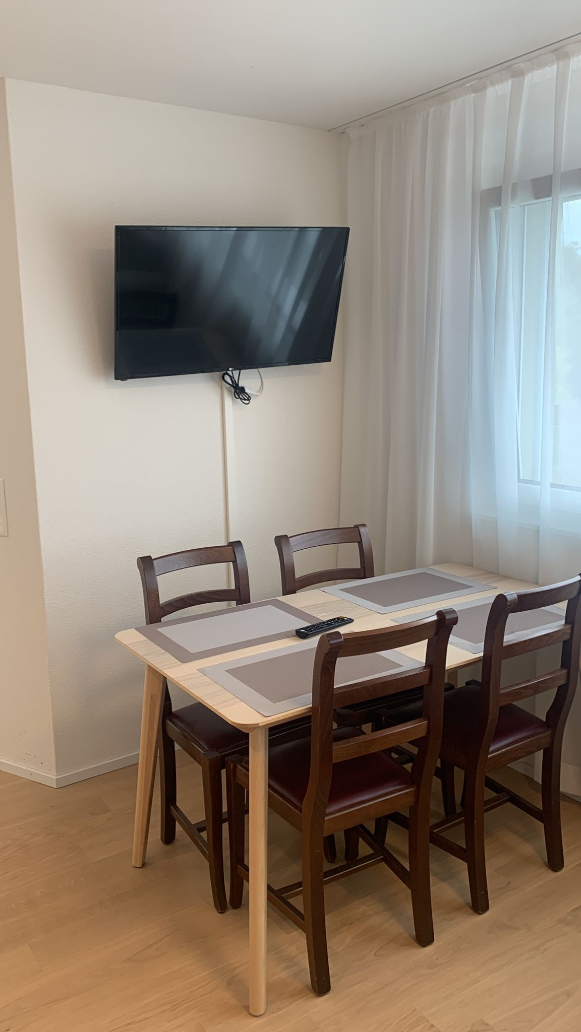 Un comedor con una mesa y sillas, un televisor de pantalla plana montado en la pared, y una ventana con cortinas de gasa.