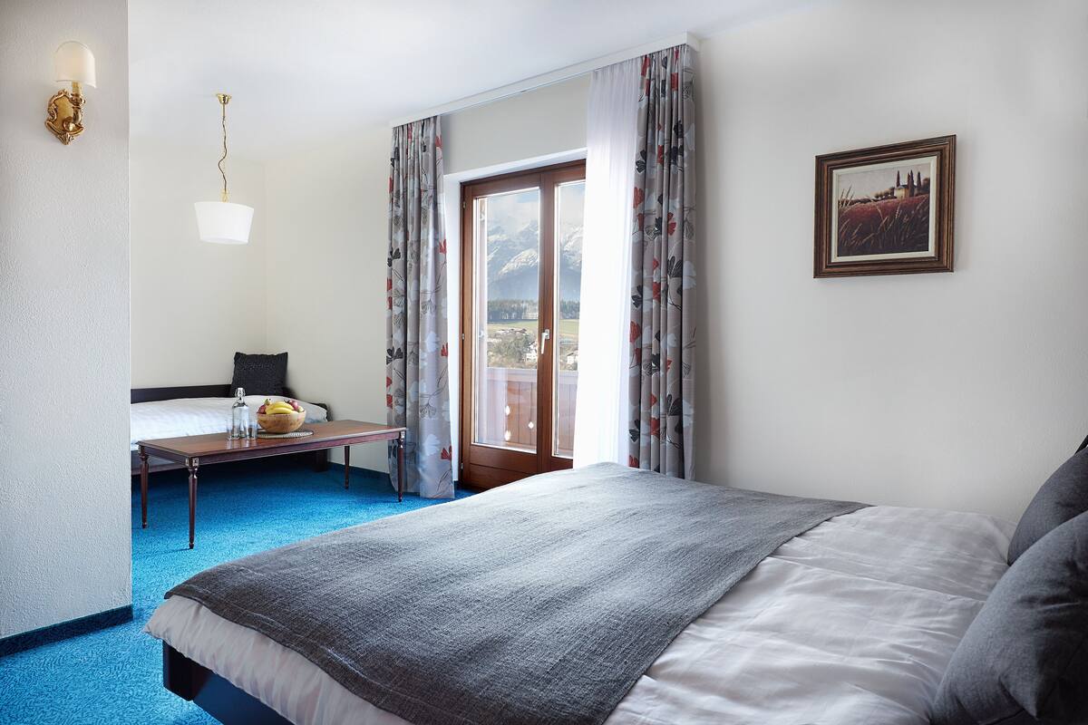 A hotel room with a bed, a small table with a bowl of fruit, a chair, and a view of the outdoors through a sliding door.