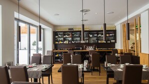 Ein Restaurant mit mehreren Tischen und Stühlen, einem Weinkühlschrank mit verschiedenen Flaschen sowie Pendelleuchten.