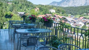 Eine Terrassengaststätte mit Metalltischen und -stühlen, umgeben von Topfpflanzen und mit Blick auf einen Berg und Häuser im Hintergrund.