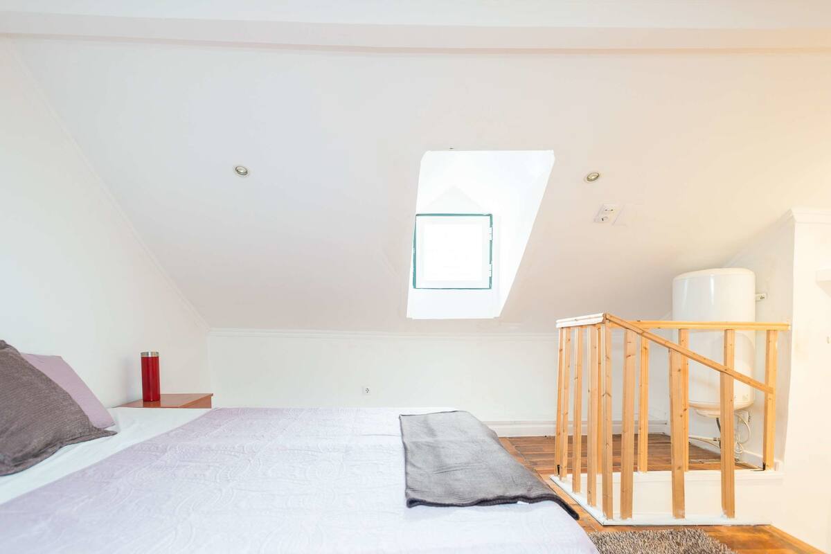 Un dormitorio con techo inclinado, tragaluz, cama con almohadas, un termo rojo y una silla de madera.
