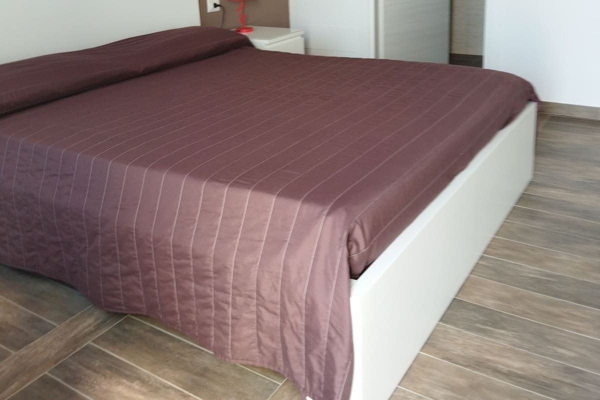 A bedroom with a large bed, a nightstand, a red lamp, and a door leading to another room.
