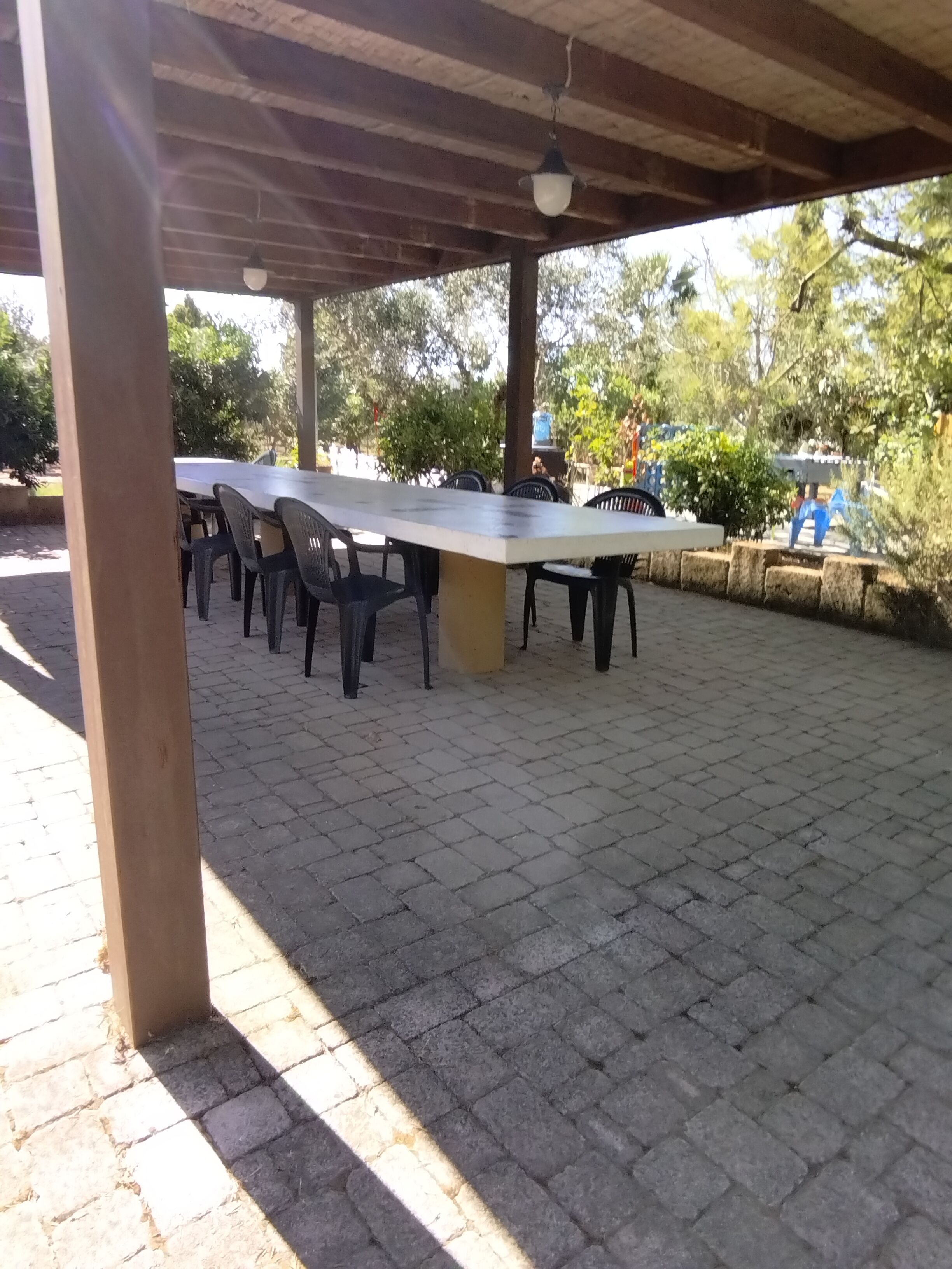 An outdoor covered patio with a long table and chairs, surrounded by stone tiles and trees.