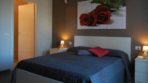 Un dormitorio con una cama, dos mesitas de noche, una mesita auxiliar, un cuadro colgado en la pared con motivos de rosas y una puerta.