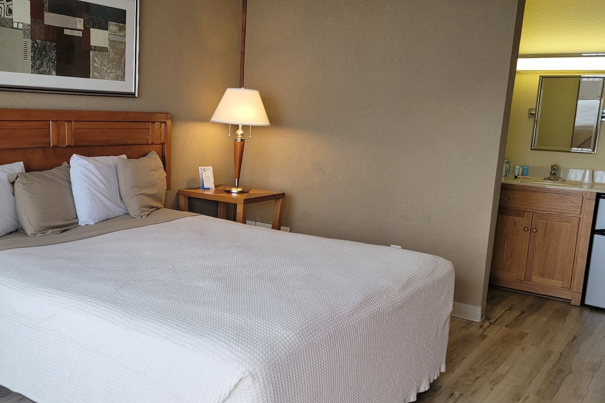 Ein Hotelzimmer mit Bett, einem Nachttisch, einer Kommode, einer Lampe und einer Küchenzeile.