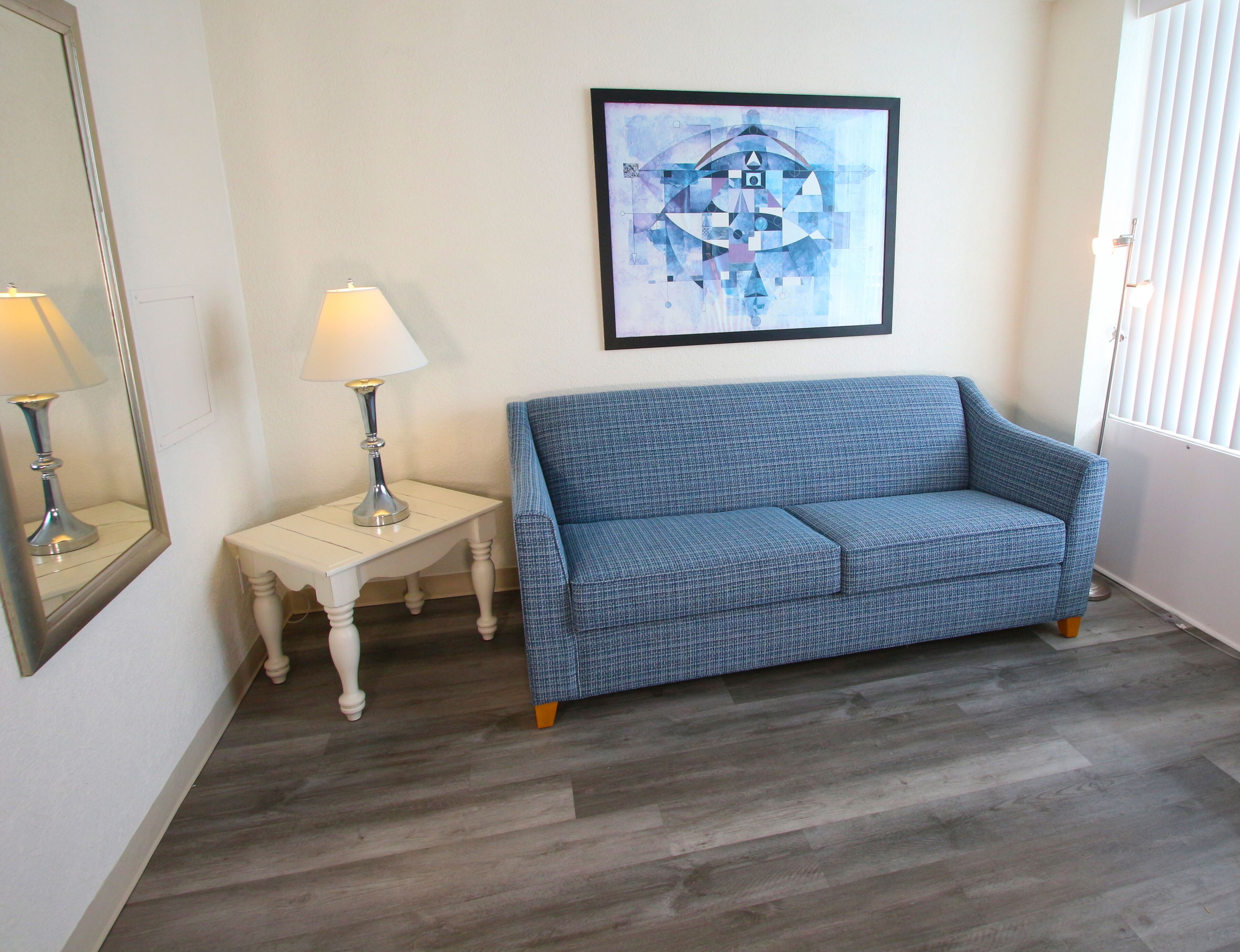 Una stanza con un divano blu dal motivo decorativo, un comodino bianco con una lampada e un quadro astratto incorniciato appeso al muro.