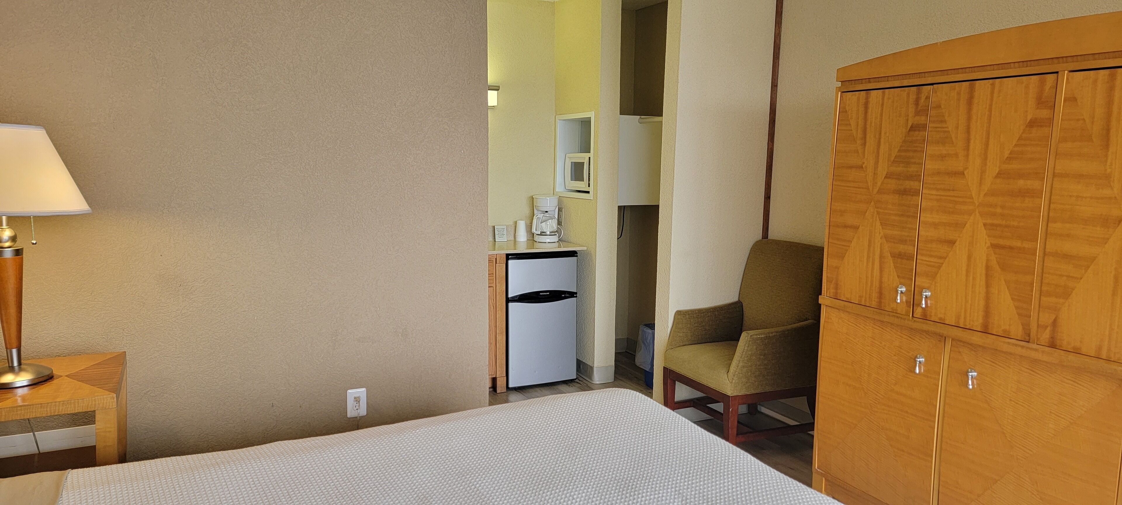 A hotel room with a bed, a wooden wardrobe, a chair, a desk with a lamp, and a kitchenette area.