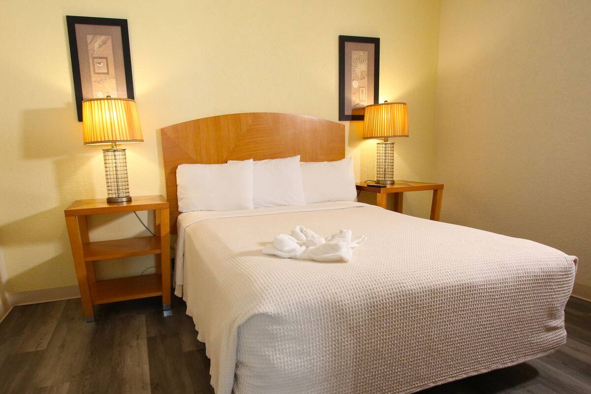 Ein ordentlich bezogenes Bett mit weißen Leinentüchern und einem schwanenförmigen Handtuch, begleitet von zwei Nachttischen mit Lampen, in einem Zimmer mit Holzböden und gerahmten Bildern an den Wänden.