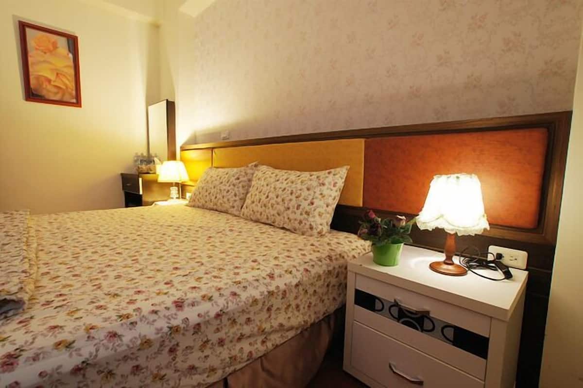 A neatly made bed with floral bedding, a bedside lamp, a small plant, and a nightstand with drawers.