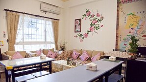A room with a long table, several chairs, a sofa with cushions, a window with curtains, and a wall with a floral decoration and a map. - The Garden Rose B&B (Taitung)