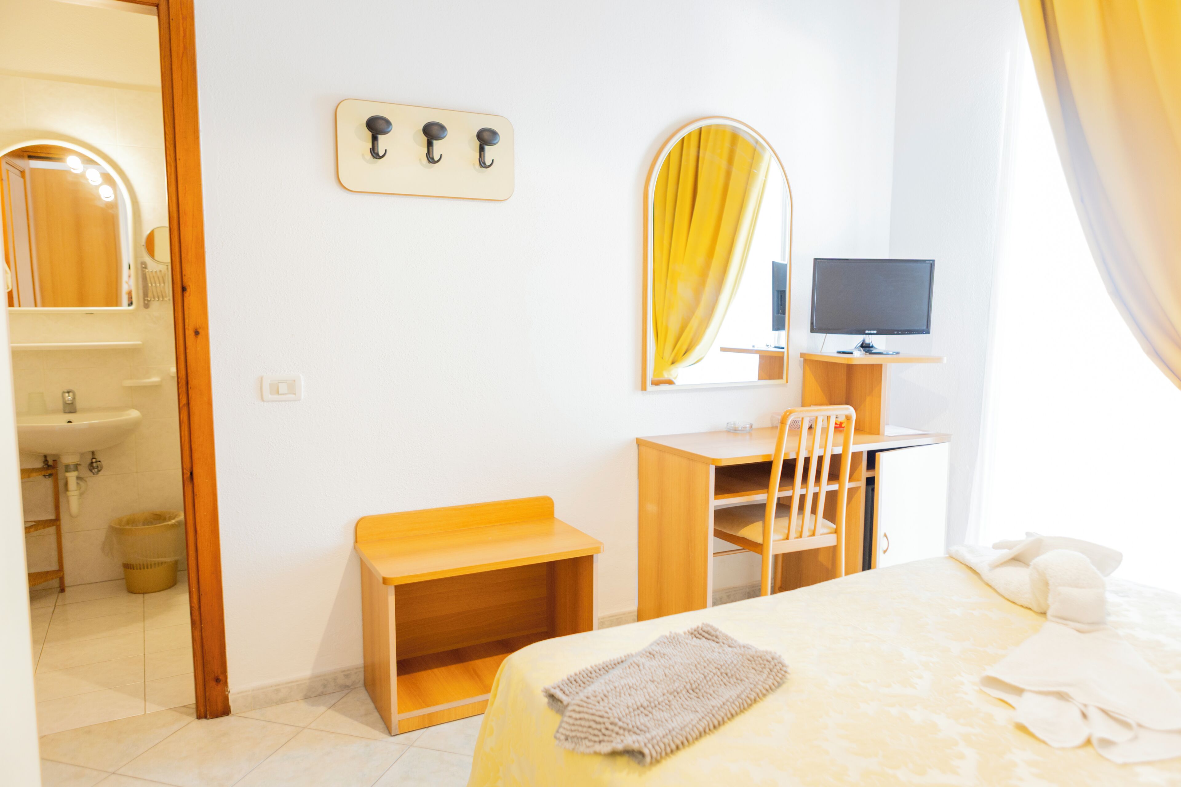 A hotel room with a bed, a desk, a chair, a television, a mirror, and a bathroom visible through an open door.