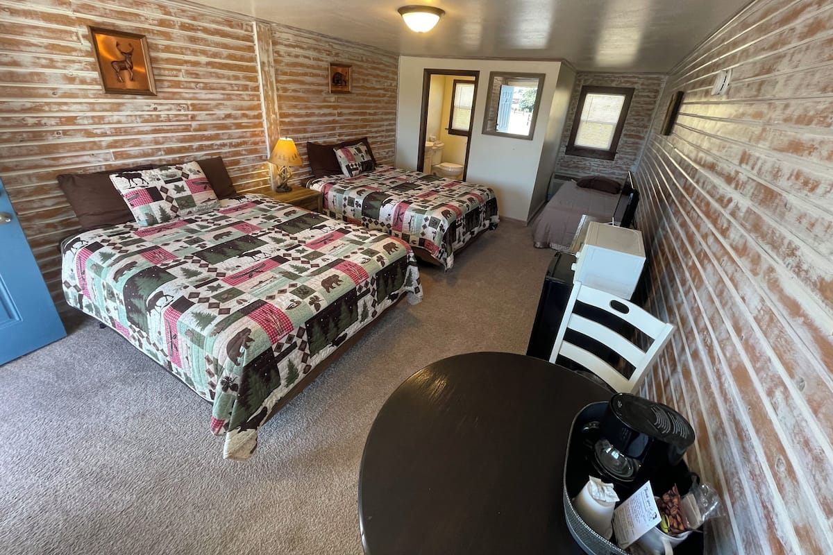 A room with two beds, a small table with a coffee maker, a trash bin, and a door leading to a bathroom.