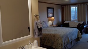 A hotel room with a large mirror, a sink with toiletries, a bed with a patterned bedspread, a bedside lamp, and a window with curtains. - El Rancho Motel (Thermopolis)