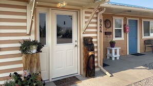A single-story building with a beige exterior, a white door, and a blue door. There is a wooden post with a carved figure next to the door, and a small white table with a potted plant on the porch. - El Rancho Motel (Thermopolis)