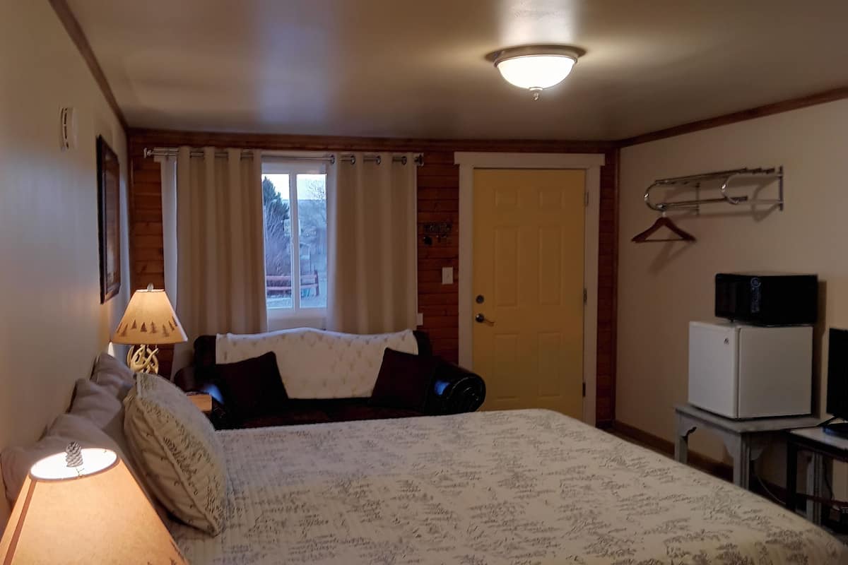 A bedroom with a bed, a bedside lamp, a window with curtains, a door, a coat rack, and a small table with a lamp.