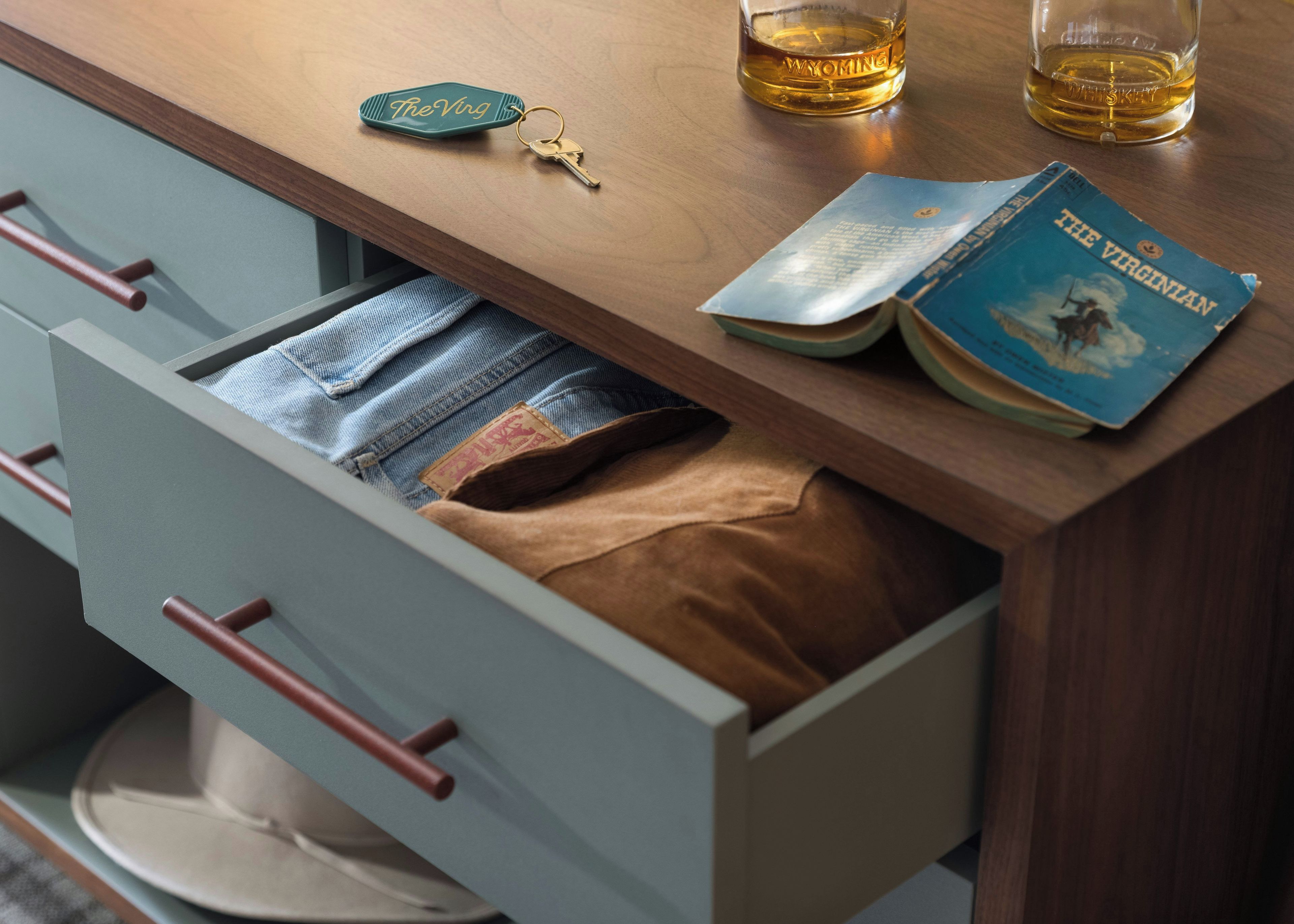 A wooden table with a book, a glass of whiskey, a keychain, and a drawer with a brown item inside.