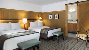 A hotel room with two beds, a bench, a chair, a wooden wardrobe, and a bathroom visible through an open door.