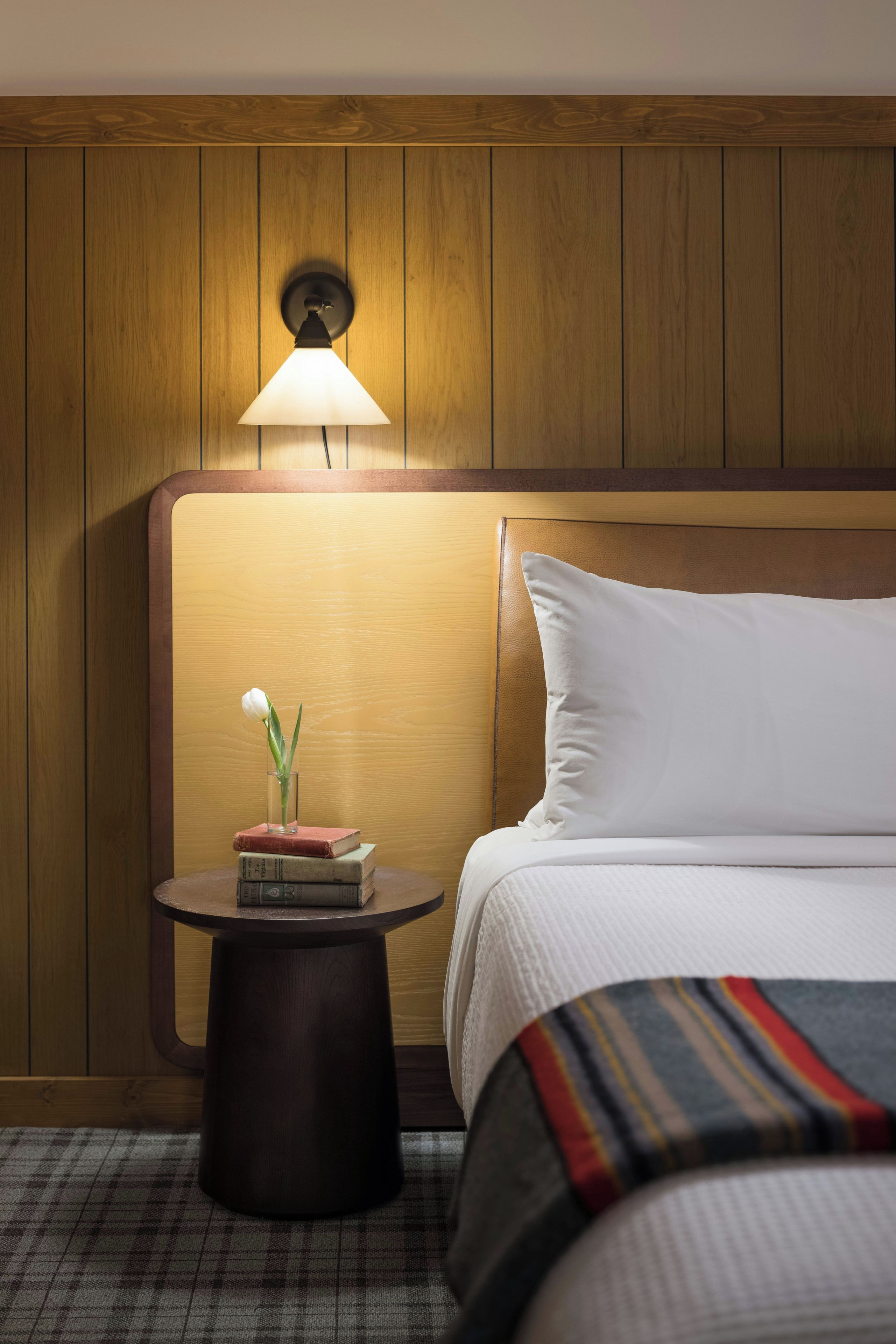 A hotel room with a wooden headboard, a bedside table with a lamp, a bed with white bedding, and a small table with a vase of flowers and books.