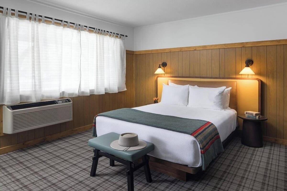 A hotel room with a bed, bedside lamps, a bedside table with a hat, a window with curtains, and a radiator.