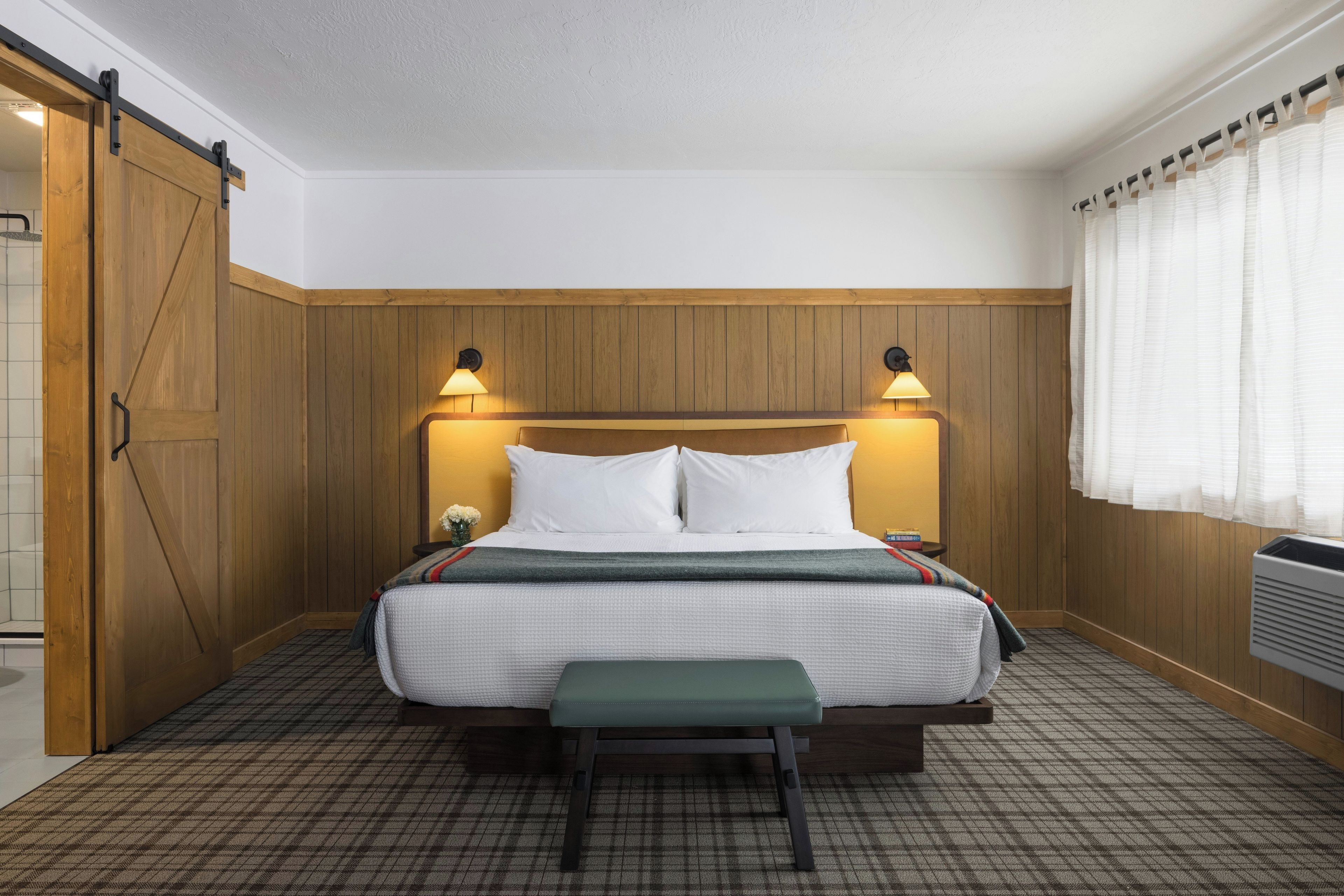 A hotel room with a wooden headboard, a bed with white bedding, two bedside lamps, a bench at the foot of the bed, and a window with curtains.