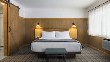 A hotel room with a wooden headboard, a bed with white bedding, two bedside lamps, a bench at the foot of the bed, and a window with curtains.