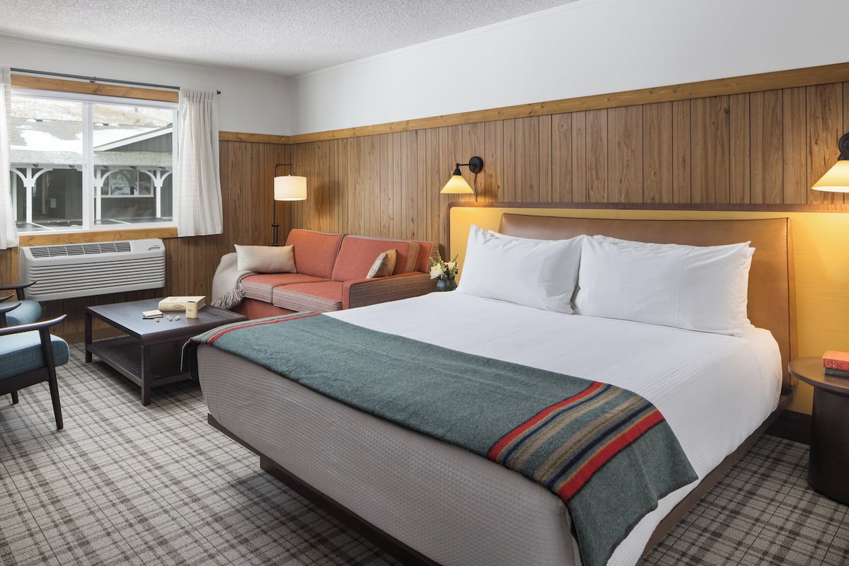 A hotel room with a large bed, a sofa, a small table, a lamp, and a window with curtains.