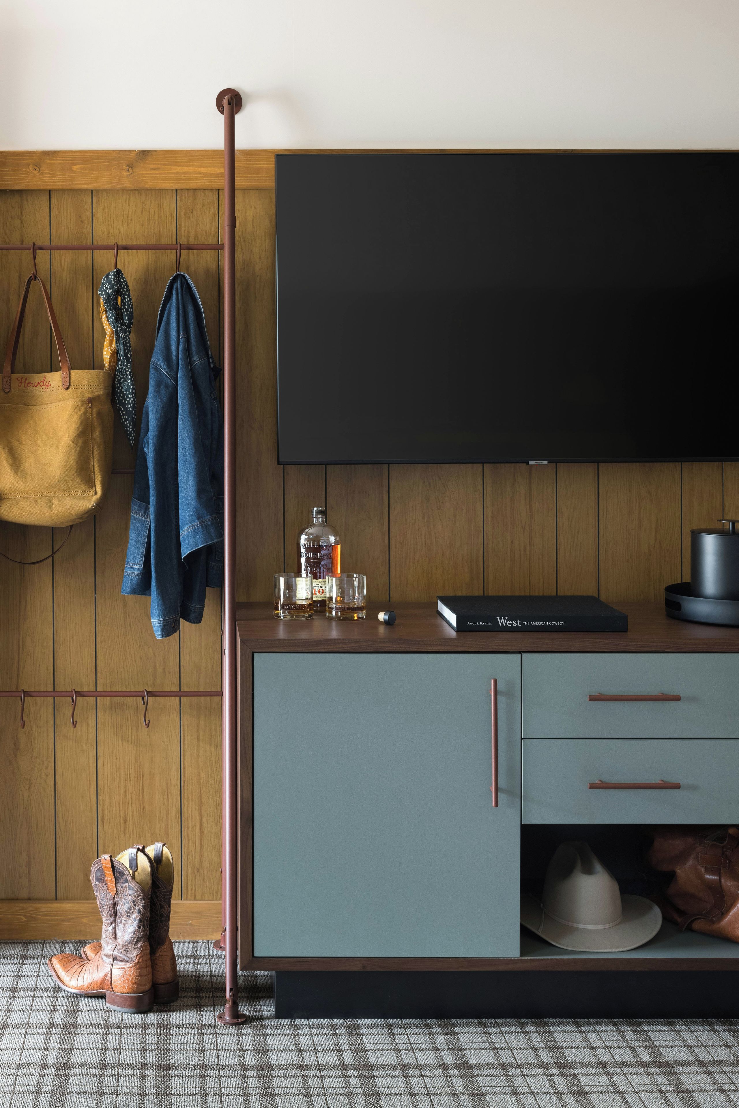 A room with a wooden wall, a flat-screen TV mounted above a wooden cabinet, a pair of boots on the floor, and a hanging coat rack with a bag and a jacket.